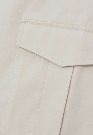 Tejido beige con un acabado texturizado, que presenta un bolsillo rectangular y en ángulo con costuras visibles y líneas limpias.