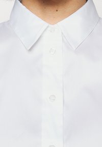 Chemise blanche à boutons avec un col classique, présentant des boutons en plastique clair et un tissu lisse et texturé. Gros plan sur la partie supérieure avant.