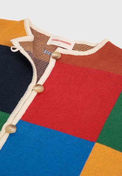 Monivärinen neulecardigan, jossa on punaisia, sinisiä, vihreitä, keltaisia ja laivastonsinisiä lohkoja. Tässä on pyöreä kaula-aukko, beige napit ja teksturoitu sisus.