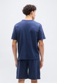 Ά νδρας με σγουρά μαλλιά, φορώντας μπλε σκούρο κοντομάνικο πουκάμισο και ταιριαστό σορτς, στέκεται με την πλάτη προς τα εμπρός σε απλό φωτεινό φόντο.