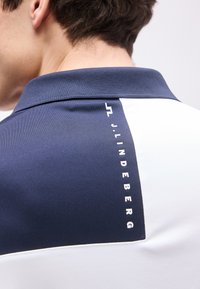 Κοντινή λήψη πίσω μέρους μπλε-ναυτικού και λευκού πουκαμίσου με κάθετο κείμενο "J. Lindeberg" και εμφανές κοντό καστανό μαλλί.