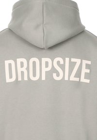 Svijetlo siva dukserica s velikim, odvažnim natpisom "DROPSIZE" u bijeloj boji na leđima. Izrađena od mekog materijala s teksturiranim završetkom.