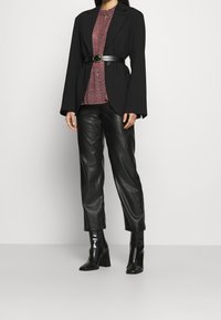 Schwarzer, oversized Blazer mit weiten Ärmeln, getragen über einem roten, gemusterten Oberteil, kombiniert mit schwarzen Lederhosen und ankle boots mit Absatz.