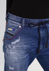 Joggers de mezclilla en azul oscuro con cintura elástica y cordón. Presentan detalles desgastados y costuras coloridas en los bolsillos.