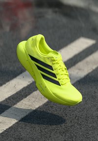 Φωτεινό κίτρινο αθλητικό παπούτσι με μαύρες ρίγες, ελαφρύ σχέδιο, υφή στην επάνω επιφάνεια και μαλακή σόλα, εμφανιζόμενο στον αέρα με φόντο έναν δρόμο.