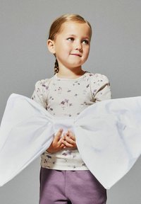 Junges Mädchen mit geflochtenem Haar, das ein blumiges Oberteil und lila Hosen trägt, hält eine übergroße weiße Schleife, die um ihre Taille gebunden ist, vor einem einfarbigen Hintergrund.