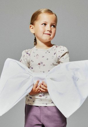 Jeune fille avec des cheveux braided portant un haut fleuri et un pantalon violet tient un grand nœud blanc attaché à sa taille sur fond uni.