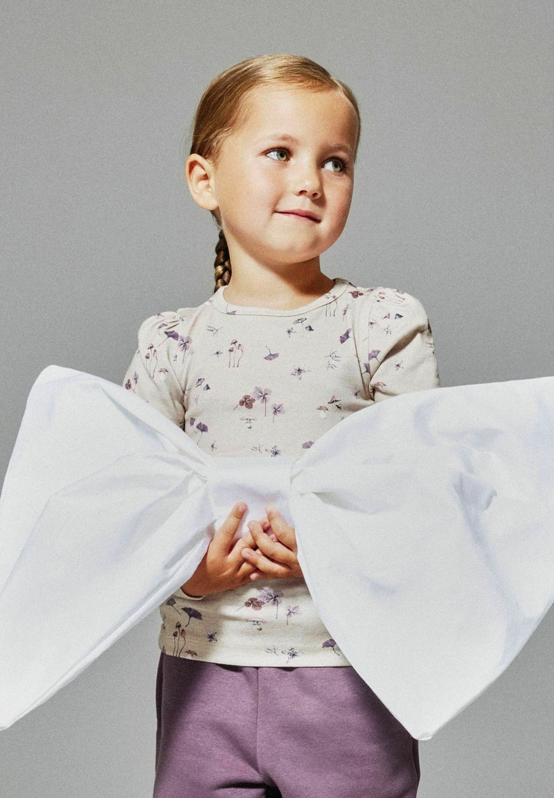 Junges Mädchen mit geflochtenem Haar, das ein blumiges Oberteil und lila Hosen trägt, hält eine übergroße weiße Schleife, die um ihre Taille gebunden ist, vor einem einfarbigen Hintergrund.