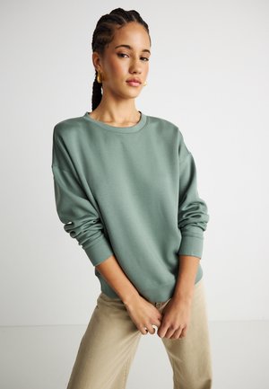 Tânără cu părul împletit, purtând cercei mari aurii, un hanorac verde cu mâneci lungi și pantaloni bej, pe un fundal simplu.