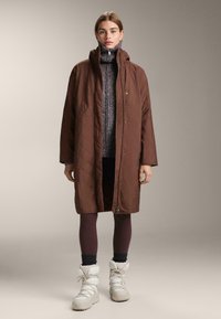 Smeđi quilted dugi kaput s kopčama, nosi se preko uzorke džempera i tamnih tajica, uparen s bijelim čizmama za snijeg.