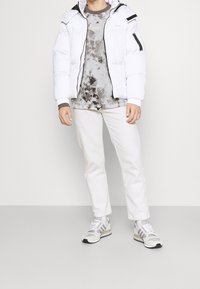 Chaqueta acolchada blanca con detalles negros, camiseta gris con efecto tie-dye, pantalones de colores claros y zapatillas grises con tres franjas y detalles texturizados.