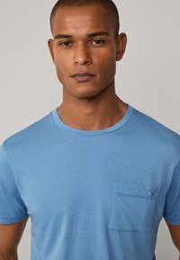 Světle modré bavlněné tričko s kulatým výstřihem, krátkými rukávy a náprsní kapsou s jemným prošíváním. Hladká textura.