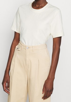 Persoon draagt een eenvoudig wit t-shirt met korte mouwen, ingestopt in hoog getailleerde beige wijde broek, staat met ontspannen armen.