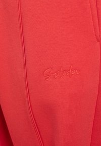 Rote Sweatpants aus weicher Baumwolle mit gesticktem "Smileden"-Logo. Das Design umfasst Seitentaschen und eine strukturierte Oberfläche.