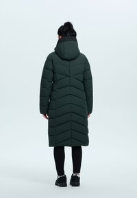 Abrigo puffer largo y verde con un patrón de chevron, capucha y cremallera; llevado sobre leggings negros y zapatos oscuros, textura suave.