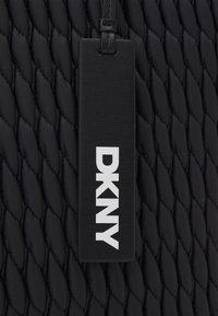 Fond en tissu matelassé noir avec une étiquette DKNY en cuir noir attachée par une fine sangle, avec des lettres blanches en gras.