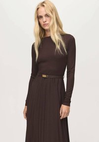 Donna bionda che indossa un vestito plissettato lungo a maniche lunghe di colore marrone scuro con una cintura sottile, in piedi contro uno sfondo chiaro e semplice.