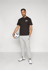 Zwart katoenen T-shirt met logo aan de voorkant, grijze joggingbroek en witte sportschoenen. De persoon houdt een voetbal met patroon vast.