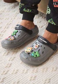 Sabots gris ornés de plusieurs breloques décoratives à l'avant, présentant différentes couleurs et formes. Bride noire et doublure en peluche pour plus de confort.