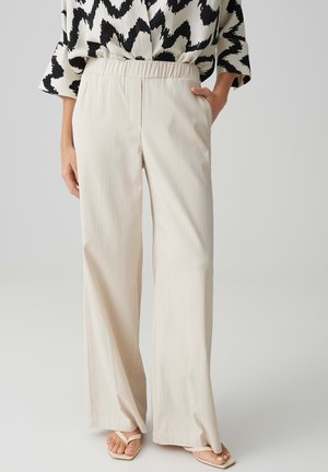 Personne portant un pantalon ample beige clair à jambes larges avec une ceinture élastique, associé à un haut à manches longues noir et blanc à motifs et des sandales blanches.