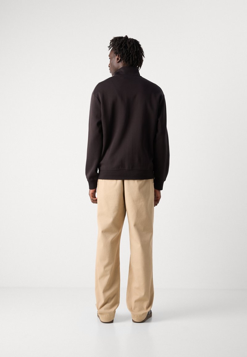 Pullover marrone scuro con collo alto, abbinato a pantaloni beige chiaro a gamba larga. Texture liscia; nessun motivo o accessorio visibile.