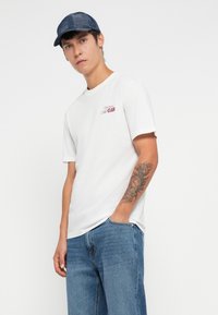Hvit bomulls t-skjorte med korte ermer, med en liten burgunder logo på venstre bryst. Kombinert med lyseblå denimjeans og en mørk caps.
