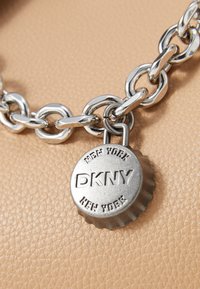 Stříbrný kovový řetěz s oválnými články a charm ve tvaru kulaté uzávěrky s vyrytým nápisem "DKNY NEW YORK", umístěný na strukturovaném béžovém pozadí.