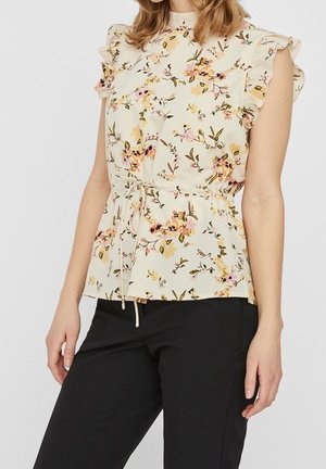Vrouw draagt een crèmekleurige bloemen top met geruite kapmouwen en een strik in de taille, gecombineerd met een zwarte broek, staand tegen een effen achtergrond.