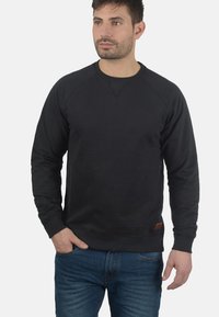 Sweatshirt noir avec des manches raglan, col rond et tissu texturé. Présente une petite étiquette en cuir en bas à gauche. Assorti avec un jean bleu.