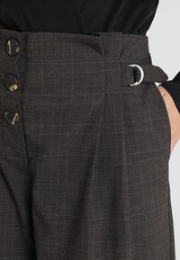 Tmavě kostkované kalhoty s vysokým pasem a opaskem, velkými knoflíky a texturovanou tkaninou. Barevná paleta zahrnuje šed�é a hnědé odstíny.