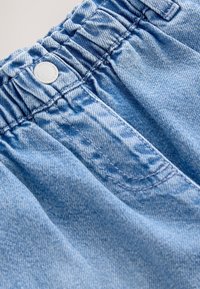 Detailní záběr světle modrých džínsů s elastickým pasem, kovovým knoflíkem a detailním prošitím v oblasti předního kapsy.