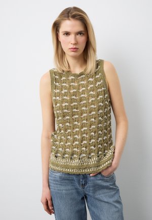 Junge Frau mit glattem blondem Haar, die ein olivgrünes, ärmelloses Stricktop und blaue Jeans trägt, vor einem schlichten hellen Hintergrund.