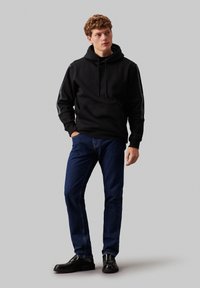 Felpa nera con cappuccio e lacci, abbinata a jeans blu scuri e scarpe nere, in piedi davanti a uno sfondo neutro.