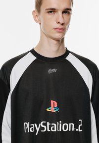 Schwarzes Trikot mit weißen Raglanärmeln, auf dem "PlayStation 2" und das Logo in Rot, Blau, Grün und Gelb abgebildet sind. Glatte Stofftextur.