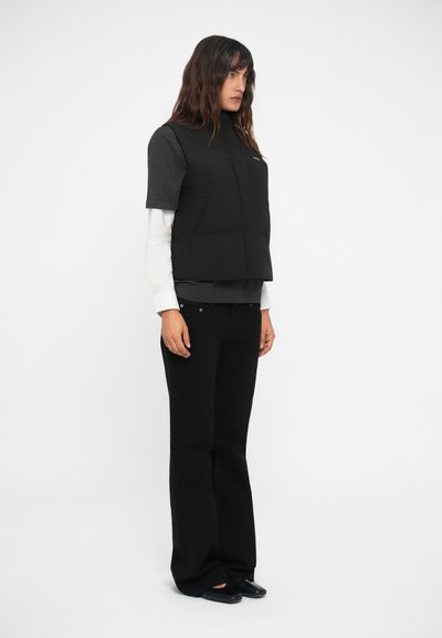 Gilet imbottito nero sopra una maglietta grigia scura, sovrapposto a una maglia bianca a maniche lunghe. Accompagnato da pantaloni neri a zampa e scarpe flat.