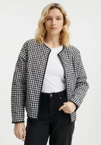 Czarna i biała kurtka w wzór houndstooth z zamkiem błyskawicznym, okrągłym dekoltem i długimi rękawami, noszona na białej bluzce i ciemnych dżinsach.