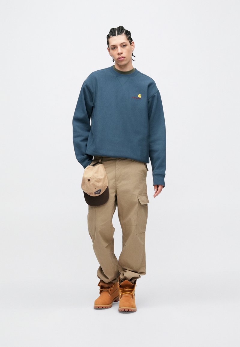 Blå sweatshirt, beige cargobukser, lysebrune støvler, holder en beige og brun kasket. Casual pasform med et logo på brystet.