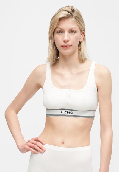 Ξανθιά γυναίκα που φοράει λευκό crop top Versace με κουμπιά και λωρίδα με λογότυπο, με το χέρι στη μέση, σε απλό γκρι φόντο.