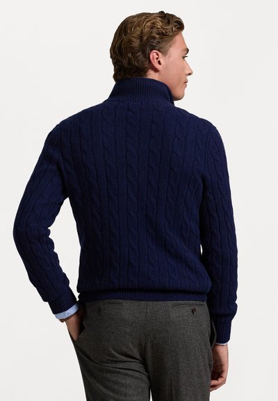 Πλεκτό πουλόβερ σε σκούρο μπλε χρώμα με πλέξη καλωδίων, που διαθέτει ψηλό γιακά και ριμπ μανσέτες. Το σχέδιο έχει υφή με ανάγλυφα, διασταυρούμενα μοτίβα.