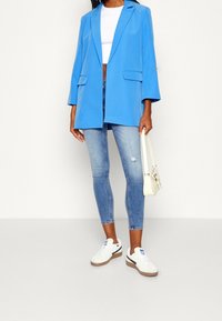 Světle modrý blazer s volným střihem a velkými předními kapsami, spárovaný s opranými skinny džíny a bílými teniskami. Drží malou krémovou kabelku.