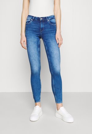 Frau in blauen Skinny-Jeans und weißen Plateausneakers, die vor einem einfarbigen hellgrauen Hintergrund steht.