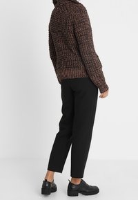 Maglione marrone lavorato a maglia con colletto alto, abbinato a pantaloni neri a sigaretta e scarpe nere, visto da dietro su uno sfondo bianco.