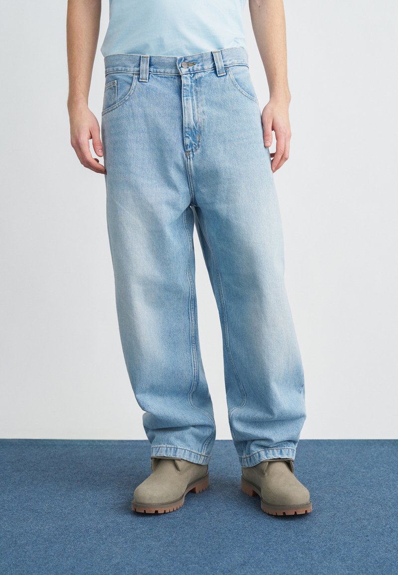 Hellblaue Jeans aus Denim mit lockerem Schnitt, fünf Taschen und Knopfverschluss, kombiniert mit grauen Stiefeln auf einem blauen Teppich.