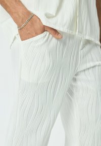 Vêtement en tissu blanc plissé avec un motif de vagues texturé. Comprend une poche et un bracelet en chaîne argenté visible au poignet.