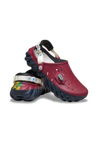 Chunky-soled klompen met kastanjebruine bovenkant, blauwe sculpted zolen, witte patronen straps en hexagonale knoopaccenten op een witte achtergrond.