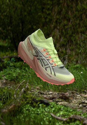 Chaussure de course vert clair et rose avec des détails noirs, suspendue au-dessus d'un sol forestier vert avec de l'herbe, de la terre et des brindilles.