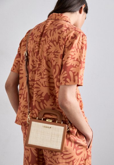 Tan handtas met een gemêleerde crèmekleurige voorkant, gouden hardware en een bruine schouderriem. Draagt crossbody, gecombineerd met een koraalkleurig patroon shirt.