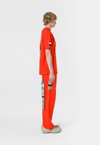 Camiseta roja y joggers a juego con detalles gráficos, presentando una textura agradable al tacto; estilizados con calzado único y un parche como accesorio en los pantalones.