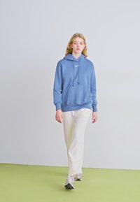 Hoodie azul de algodão com um bolso frontal e logotipo branco, combinado com calças bege claras e ténis cinzentos, numa fundo neutro.