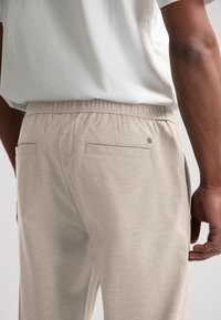 Lys beige joggebukse med elastisk midjebånd, to baklommer og en glatt tekstur. Minimalistisk design med liten logodetalj.